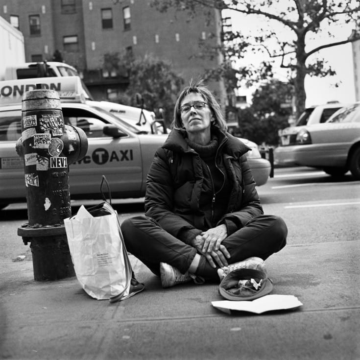 Woman sitting on sidewalk in front of fire hydrant, begging for change.