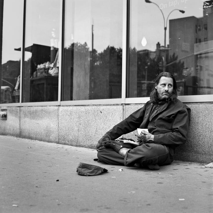 Man sitting in front of whole foods in zazen posture with hat on ground, begging for changing.