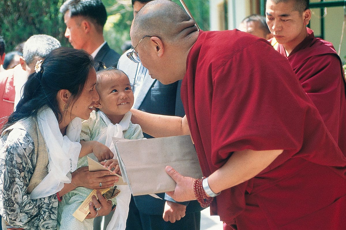 Dalai Lama wearing red robes holding the head of a young child. There are people in the background and the child is smiling.