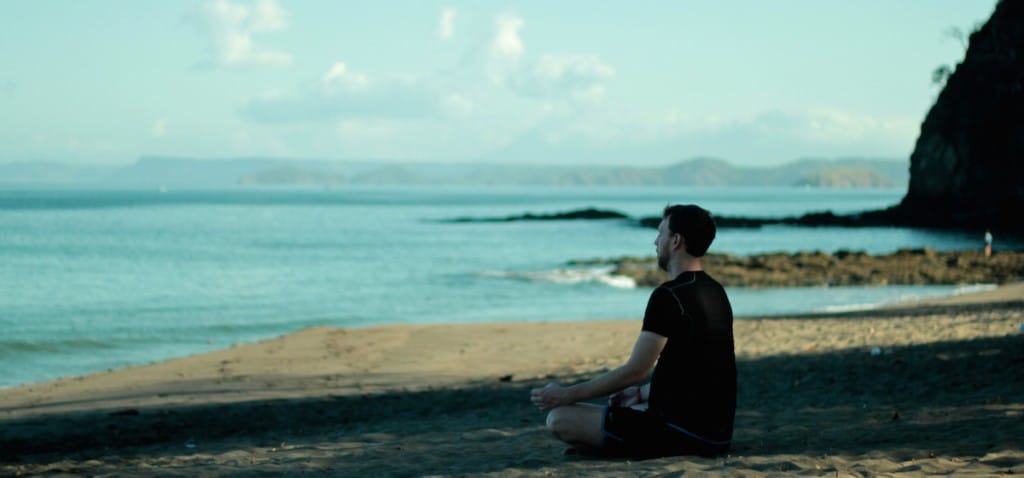 A man meditates on a beach. He is wearing a black t shirt and black shorts, and is looking out on the ocean.