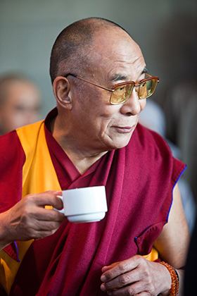 The Dalai Lama holding a tea cup. He is wearing red robes and an orange sash and clear glasses.