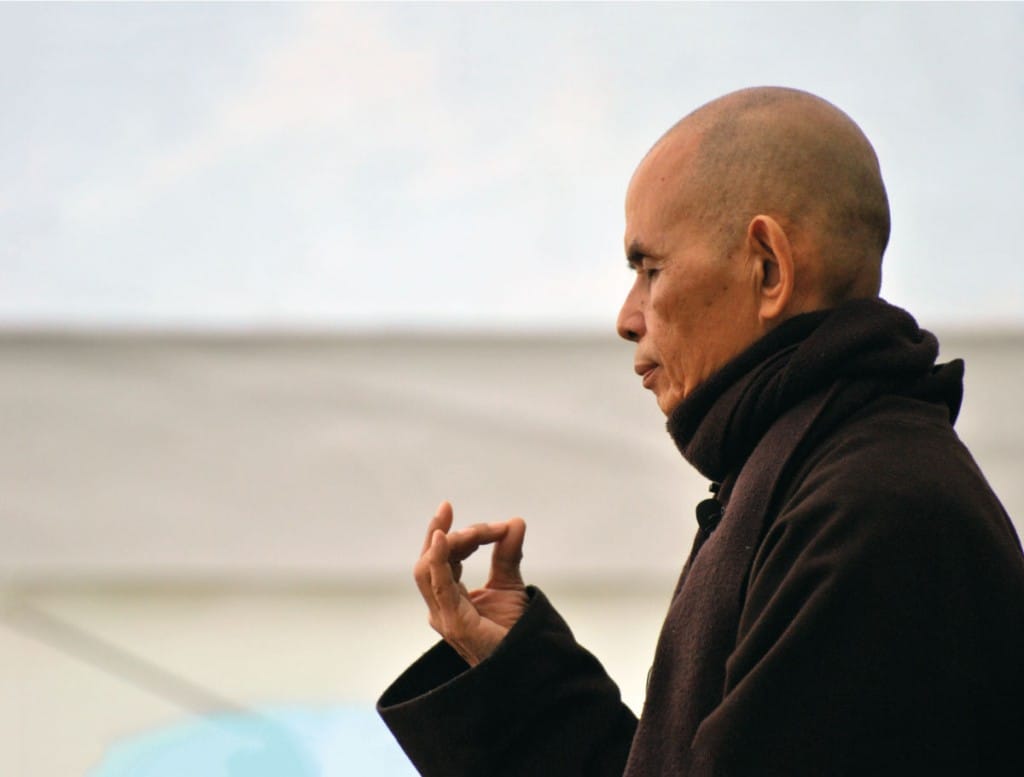 thich nhat hanh. He has on brown robes and is holding his thumb and pointer finger together.