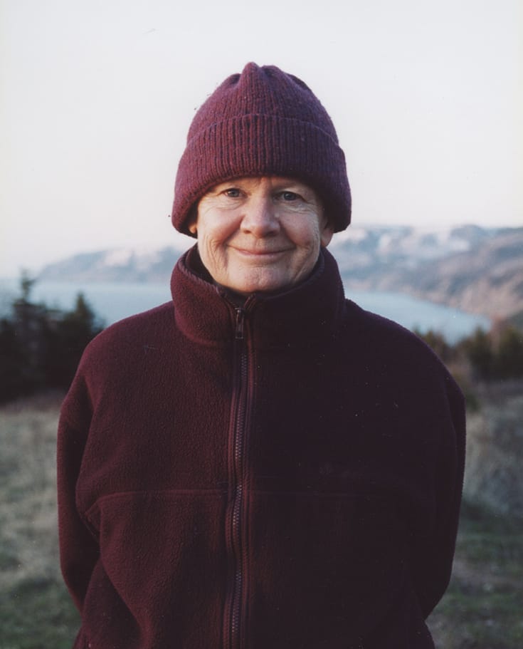 Pema Chodron smiling at the camera. She is wearing a purple sweater and winter hat. She is standing in front of a cliff near a field.