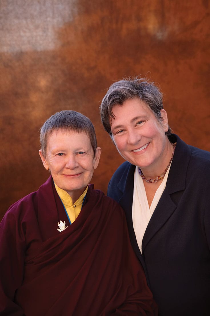 Pema Chodron and k.d. lang. Pema Chodron is wearing maroon robes with a mustard yellow shirt underneath. k.d. lang is wearing a black blazer over a white v-neck shirt. They are both smiling.