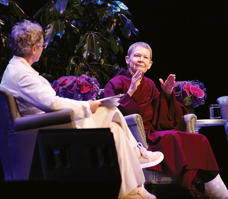 Two people sitting in armchairs having a conversation. The one on the left is wearing all white clothing and running sneakers, with short hair and clear glasses. The one on the right is wearing maroon robes with a mustard yellow shirt underneath. 