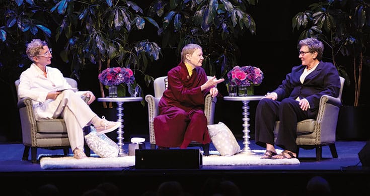 Three people on a stage sitting in armchairs with tables in between them with flowers on the tables. The person on the left is wearing all white, the person in the middle is wearing maroon robes with a mustard yellow shirt underneath, and the person on the right is wearing dress pants and a blazer with sandals.