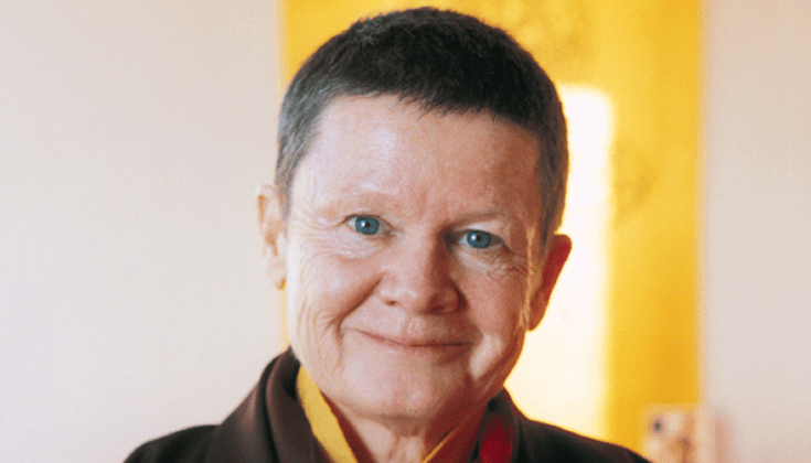 Pema Chodron smiles at the camera. She has bright eyes and short brown hair, and is wearing maroon robes with a mustard yellow shirt underneath.