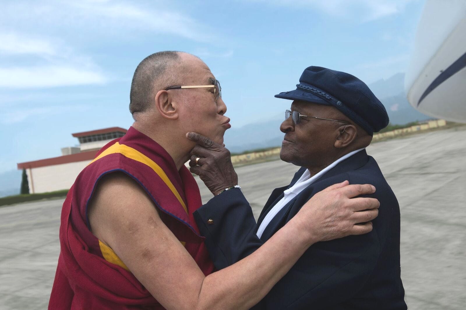 O Dalai Lama e o Arcebispo Desmond Tutu.  O Dalai Lama está vestindo uma túnica marrom com uma faixa amarelo mostarda.  Desmond Tutu usa terno e chapéu preto.