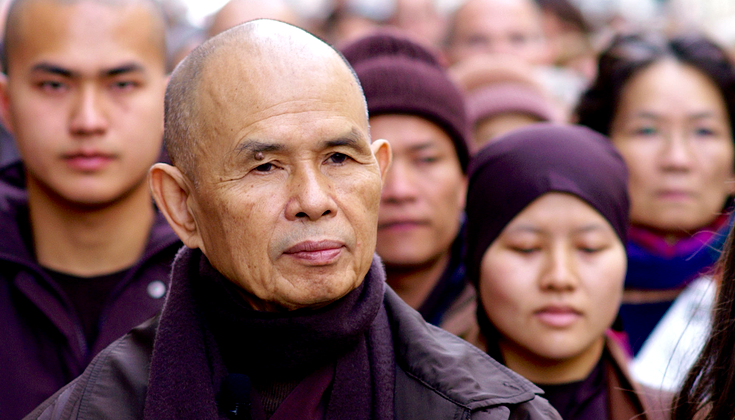 A bald man in purple robes looks off into the distance. He is in a crowd of people, all wearing purple.