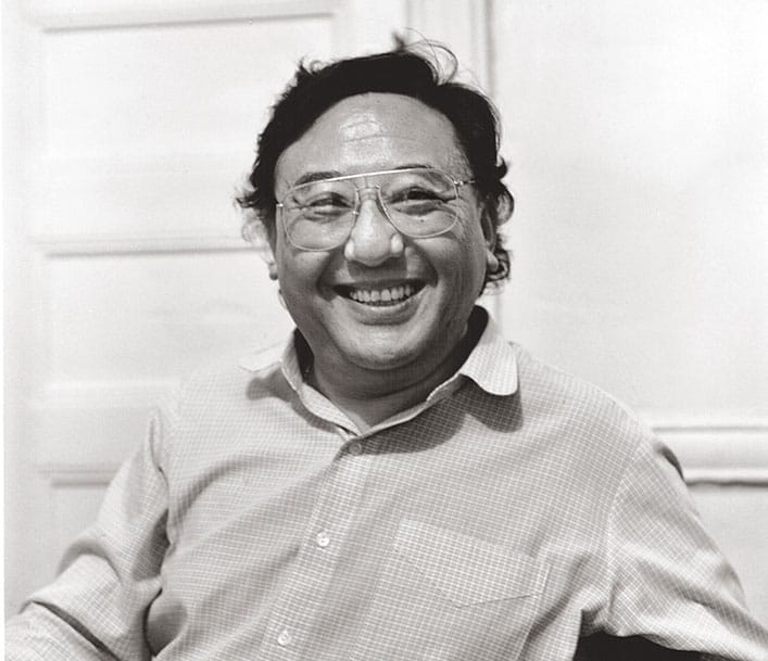 A black and white photo of a man smiling. He is wearing a checkered button up shirt and glasses.
