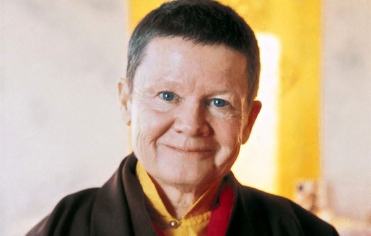 Pema Chodron smiling at the camera. She has short hair and bright eyes, and is wearing brown coloured robes over a mustard yellow shirt.