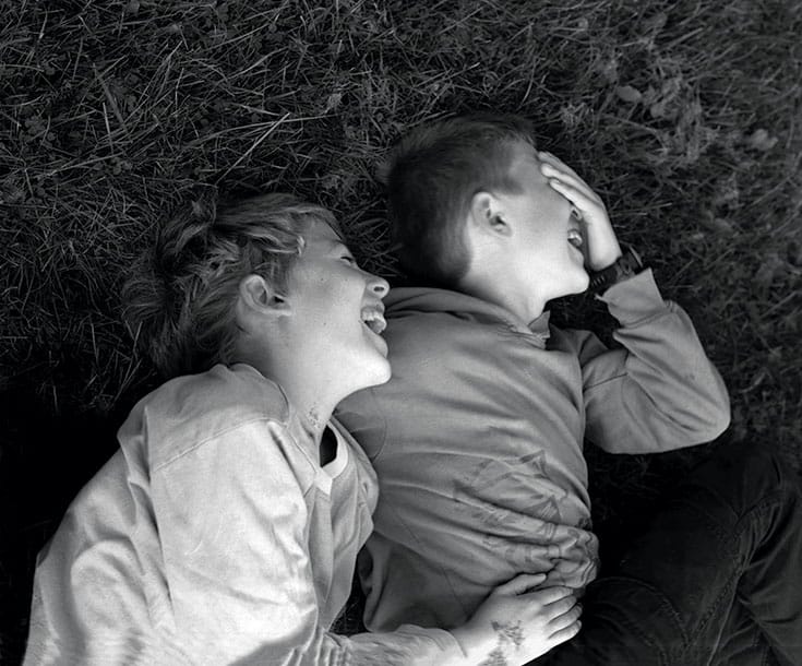 Black and white photo of two young children hugging and laughing in the grass.