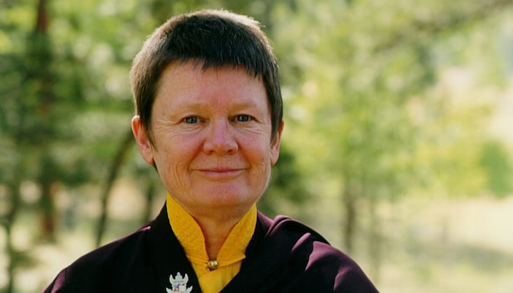 Pema Chodron smiling at the camera. She has short brown hair and is wearing maroon robes over a mustard yellow shirt. There are trees in the background.