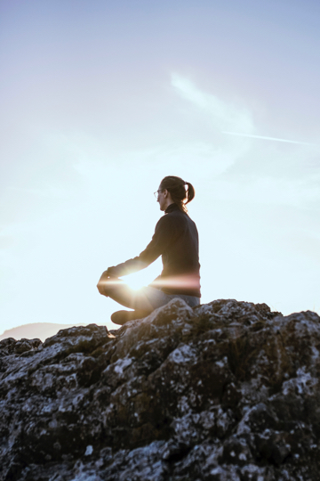 A woman sits with her legs crossed on a rock. She is meditating. She is wearing pants and a long sleeve shirt with her hair in a ponytail. The sun is bright and peeking through her arms.