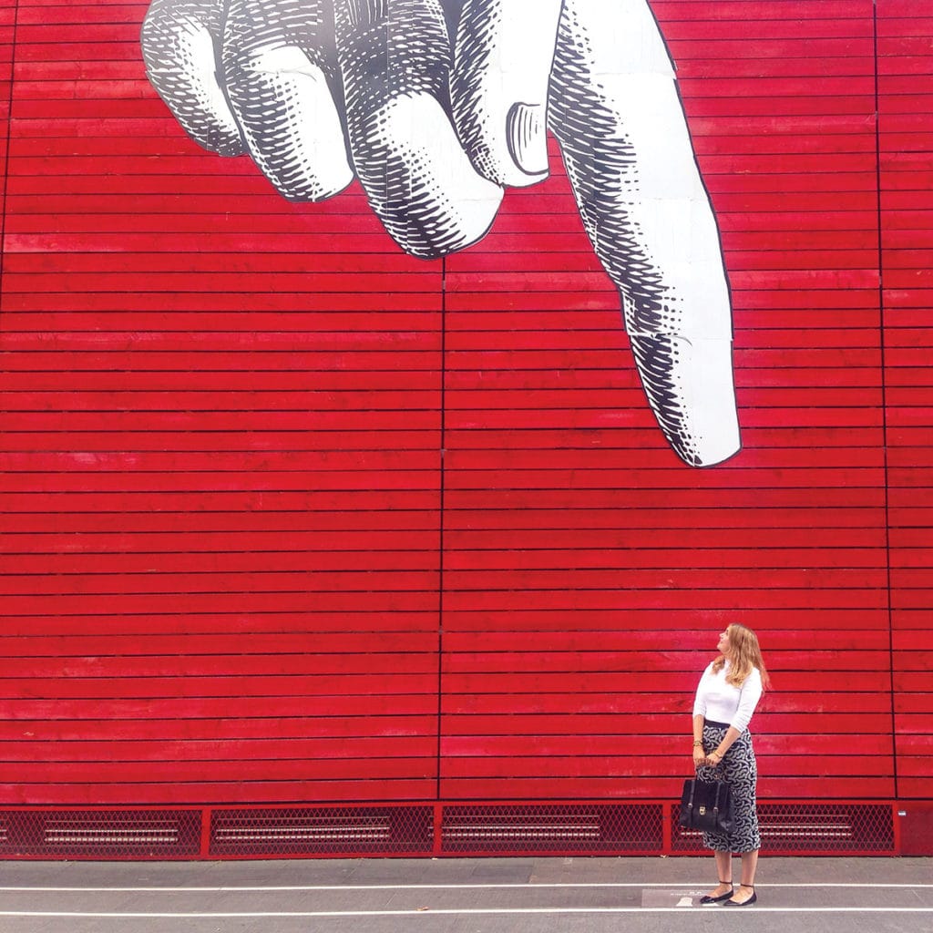 Street art of a finger pointing down at the ground. A woman stands under it.