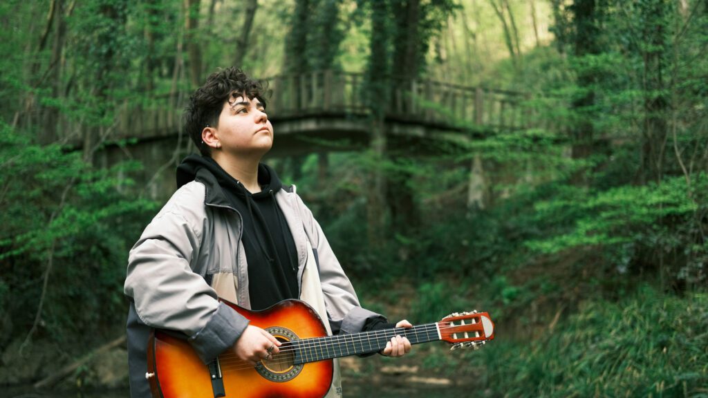 A musician holding a guitar in the woods, listening.