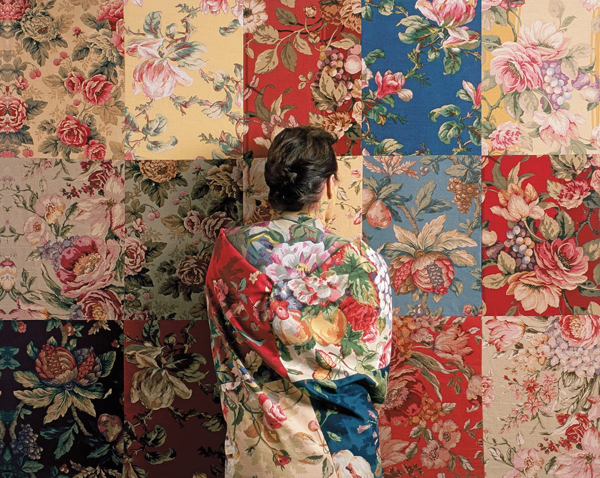 A woman standing in front of a quilt made out of floral patterns. She is draped in the same quilt.