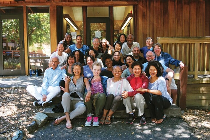 A group of people sitting outside a wooden cabin. Everyone is smiling.