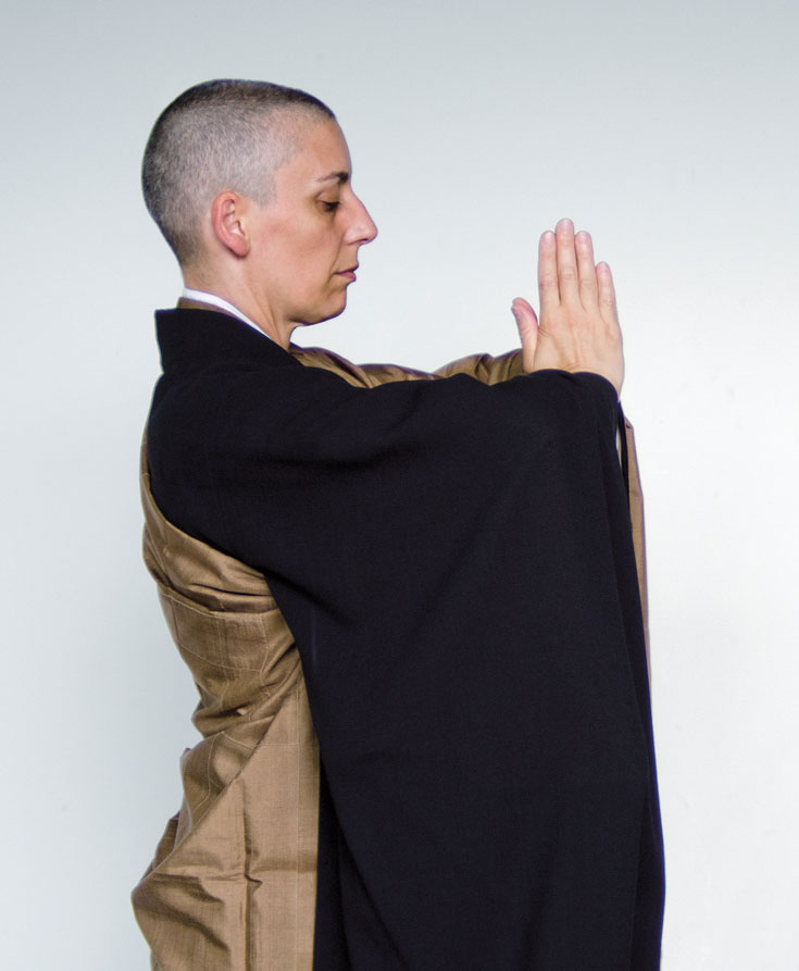 A person stands in monks robes with their hands pressed together in front of their chest.