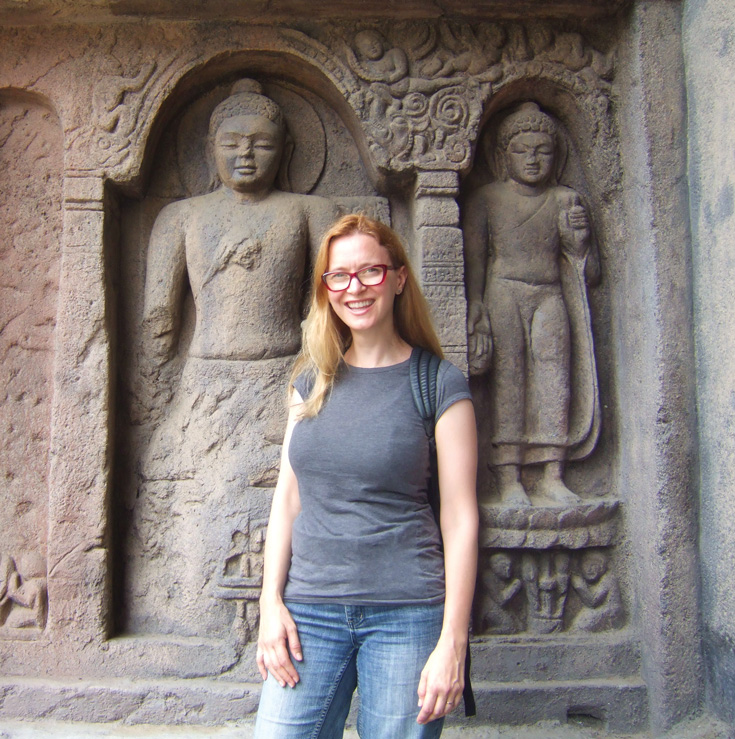 A woman standing in front of ruins that have the Buddha carved out in them. She is wearing red glasses, grey shirt, and jeans.