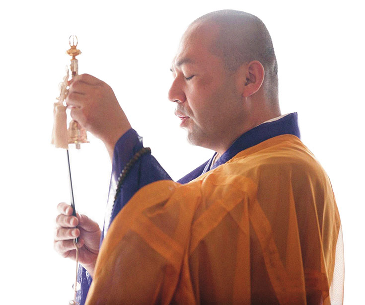 A man stands with his eyes closed, wearing orange monk robes.