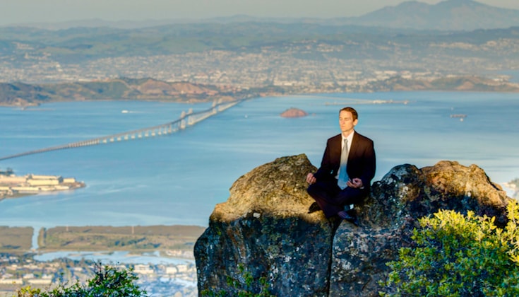Man in a business suit sitting on a hilltop, meditating, with city skyline in background.