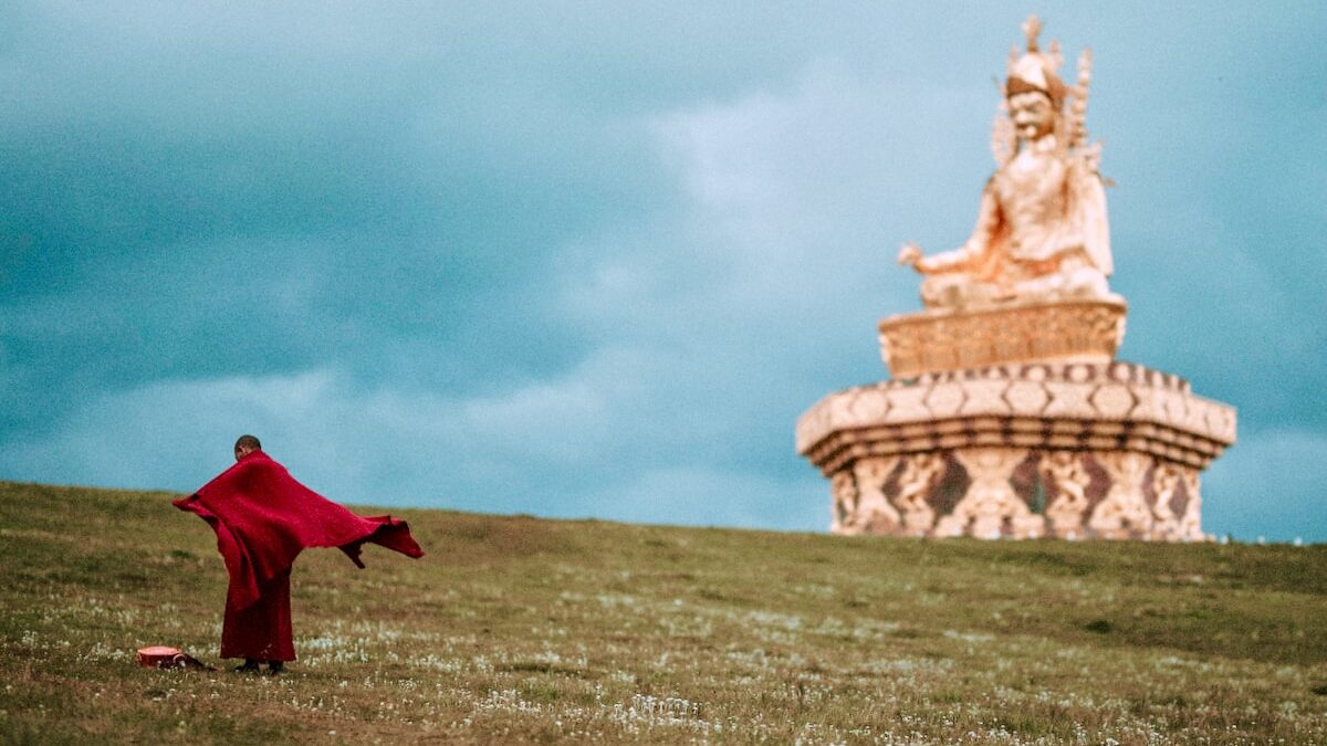 A monk standing in a field, with a statue of Padmasambhava in the distance.