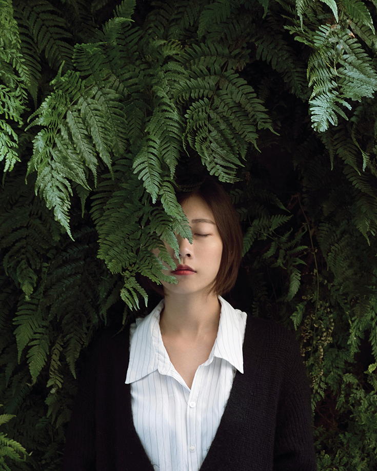 A woman wearing a white shirt under a black cardigan. Half her face is covered with a leaf.