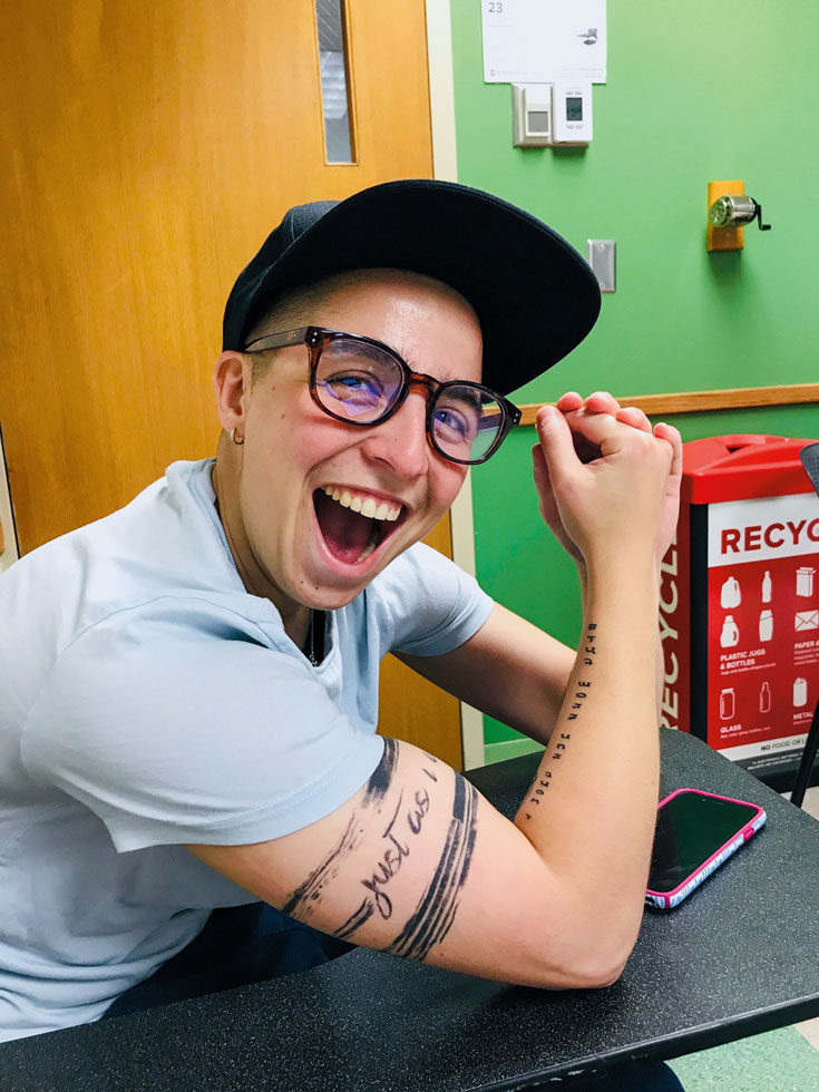 A photo of a person smiling. They are wearing a hat and glasses and have their right arm on a table with a tattoo showing.