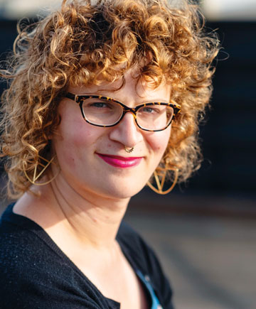 A person smiling at the camera. They have curly hair, glasses, nose ring and bright lipstick.