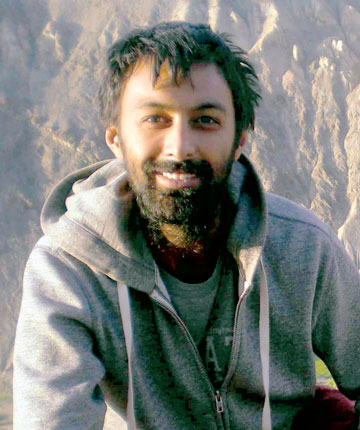 A man smiling in a grey hoodie against a nature backdrop.