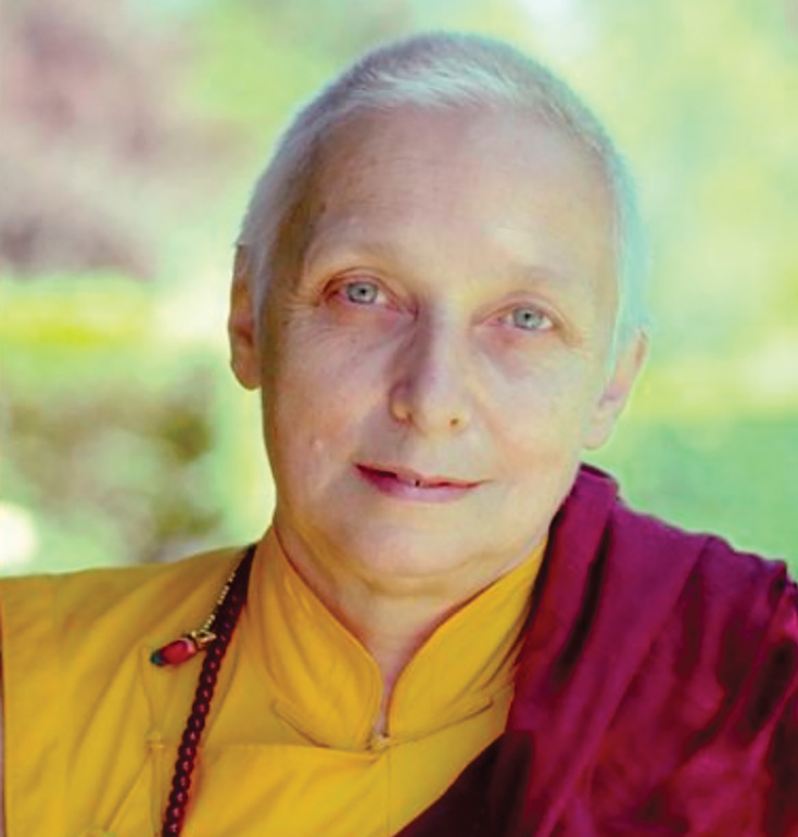 A woman with short hair smiles at the camera. She is wearing a mustard yellow shirt with a red sash over one shoulder.