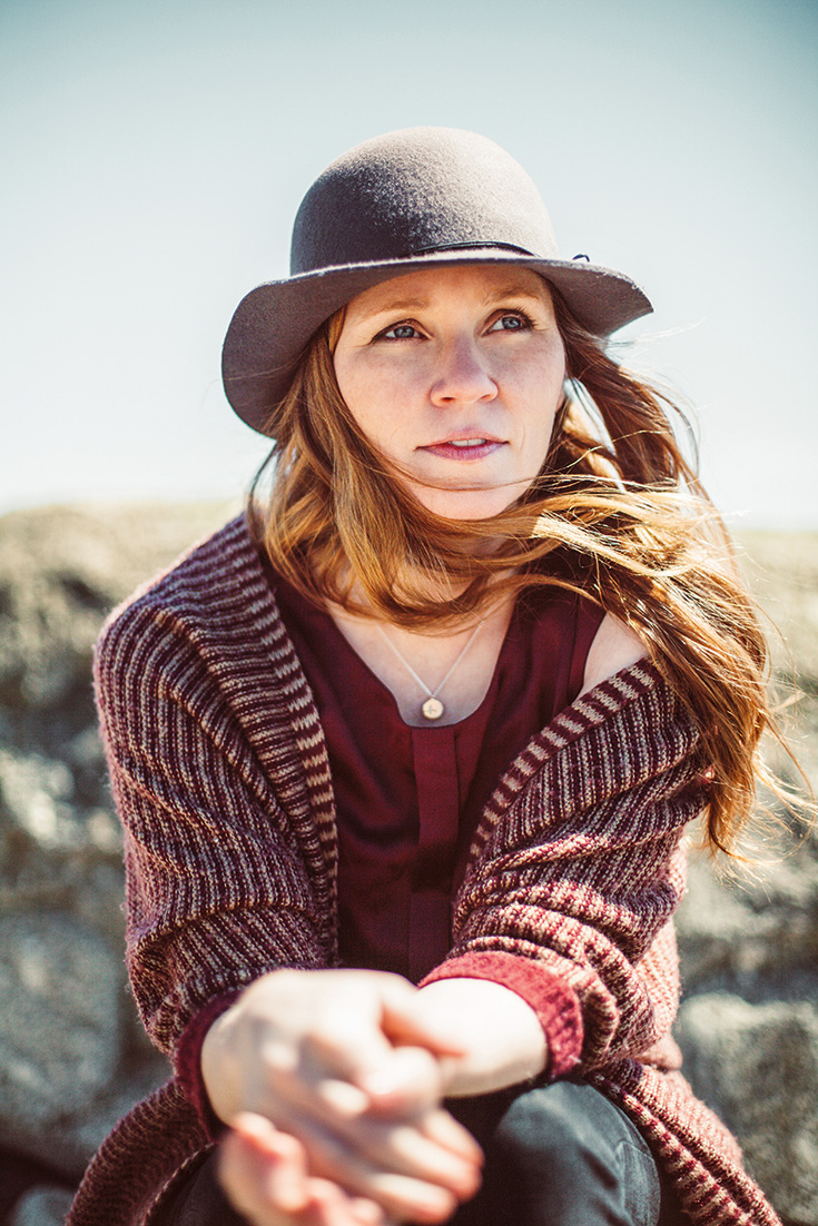 A woman wears a grey hat. She has red hair and is wearing a red tank under a red striped sweater.