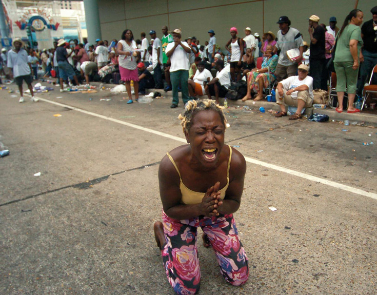 A woman with floral pants and a yellow tank top leans on her knees. Her hands are in a praying position, and she is screaming.