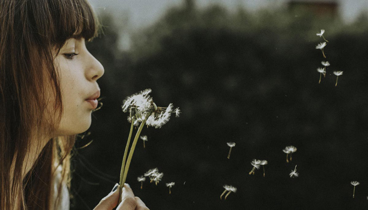Woman with brown hair and bangs blows on a dandelion.