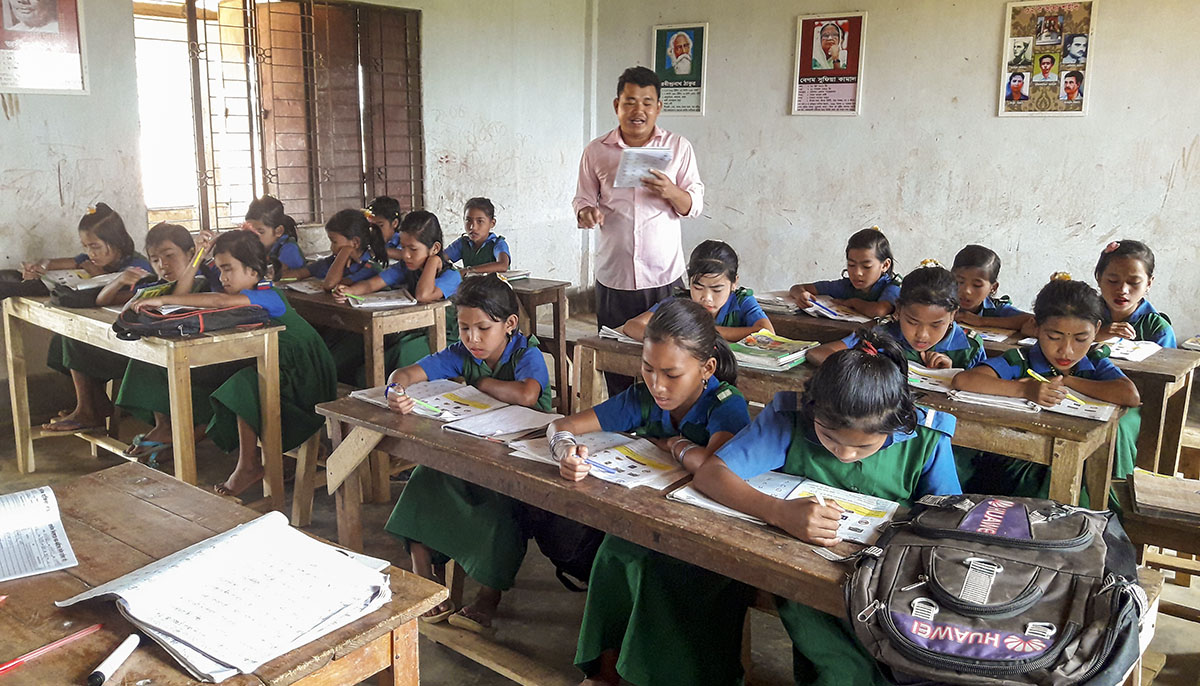 A classroom with all young girls. They are all wearing blue and green uniforms. A man is teaching.