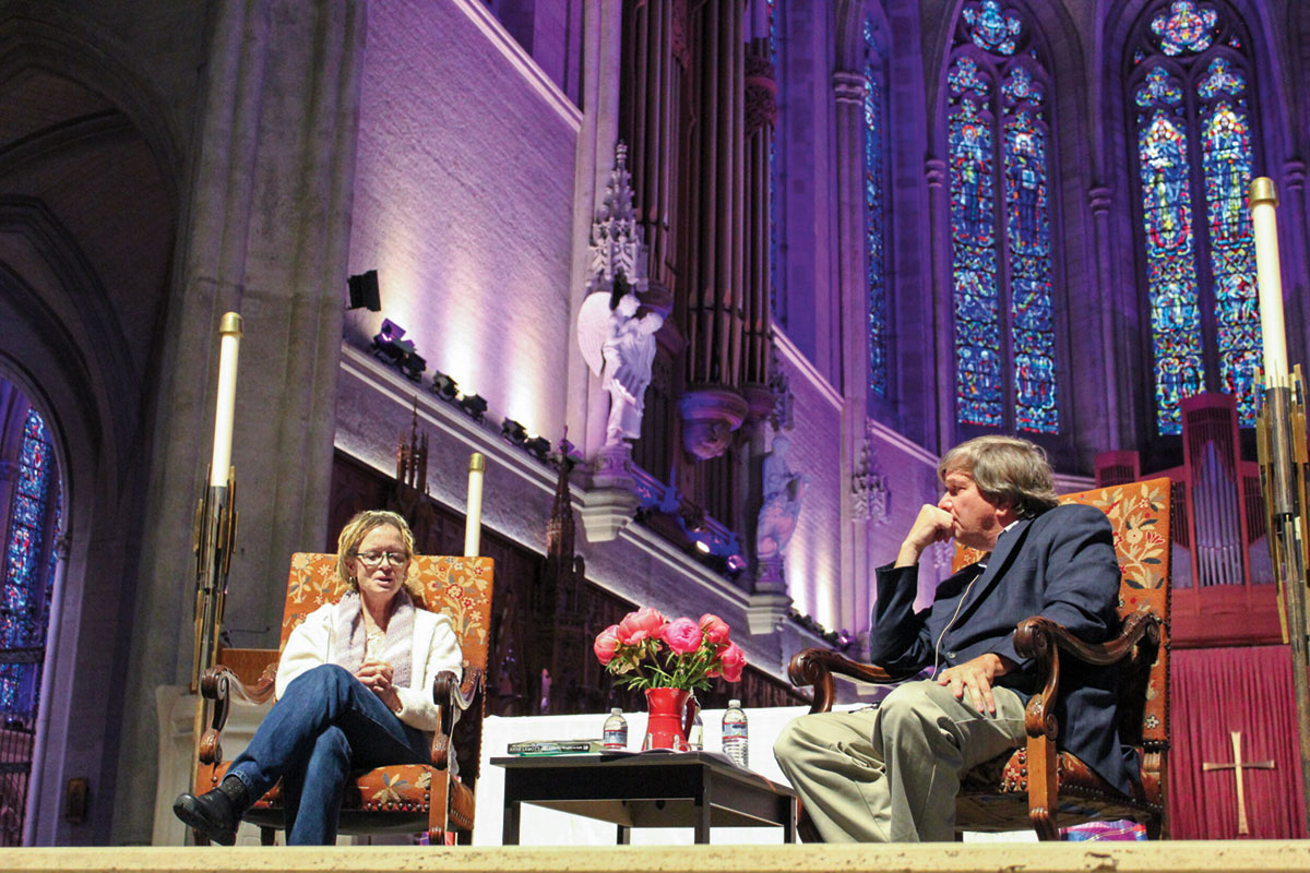Two people on a stage. There are high ceilings and stained glass windows in the background.