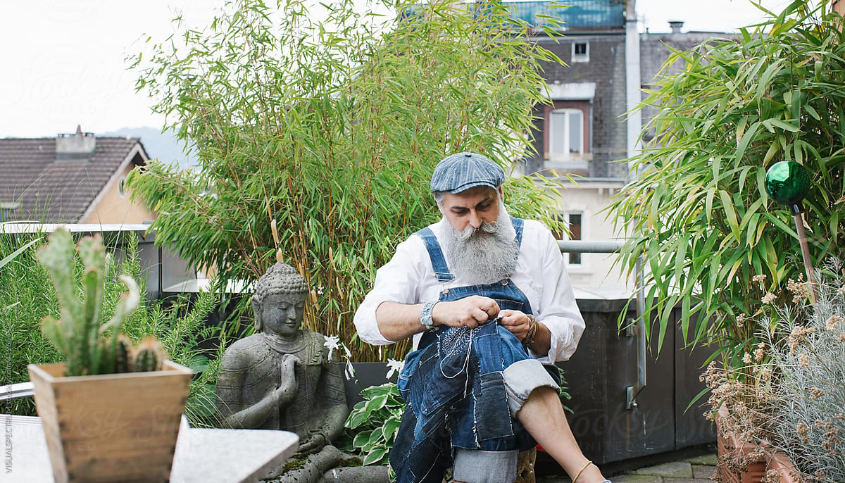 A man sewing. He is wearing denim overalls and a denim hat. He is surrounded by plants.