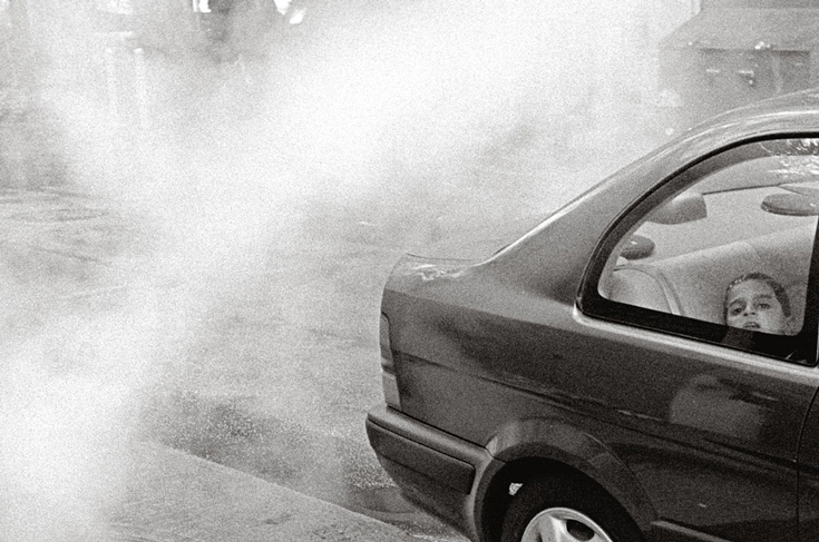 A black and white photo of the back of a car. There is a young child's face peeking at the window of the backseat.