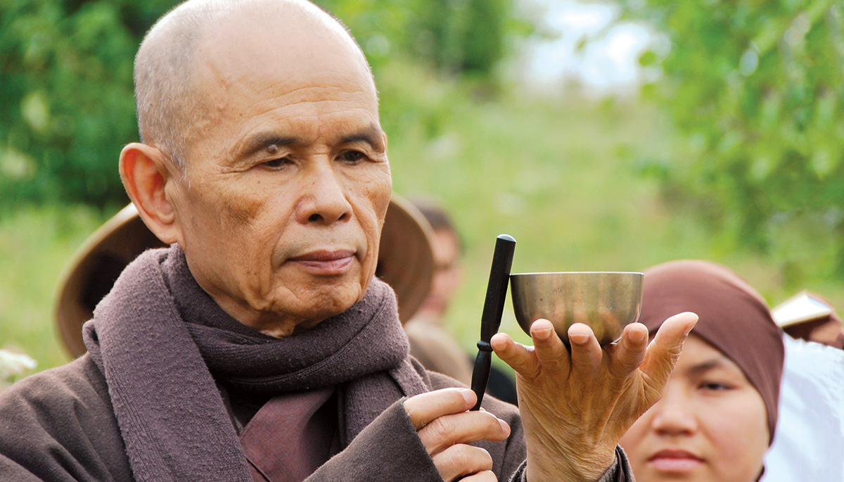 A man holding a small bowl. He is wearing purple robes with a purple scarf.