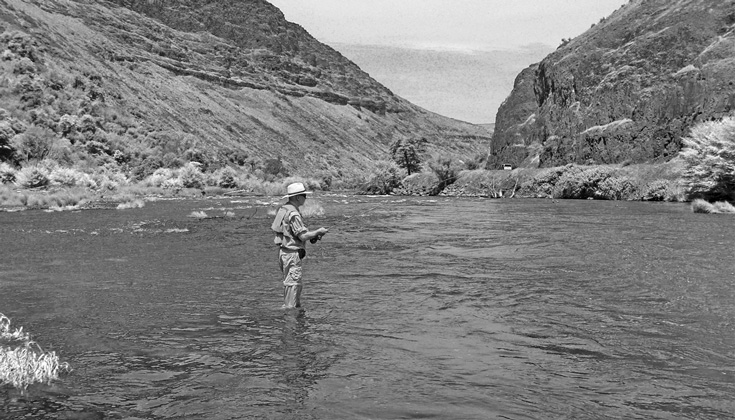 A black and white photo of a man in a body of water with cliffs in the background.