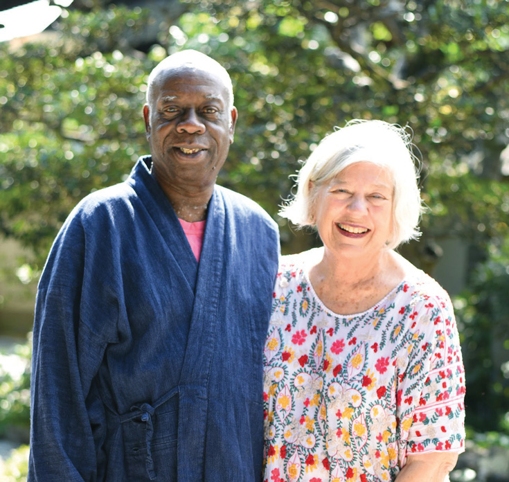 A man stands with his arm aroound a woman. The man is wearing a blue robe and the woman is wearing a patterned shirt. Trees are in the background.