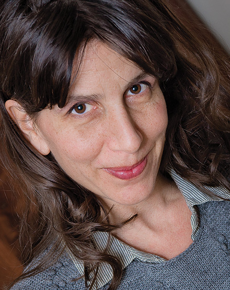 A woman with long brown hair and bangs smiles. She is wearing a striped collared shirt over a grey sweater.