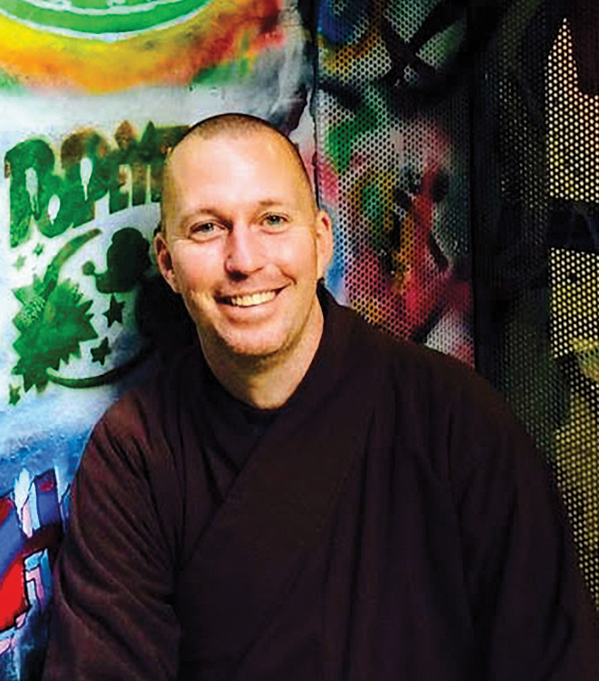 A man smiles in purple robes. There is a spray painted wall in the background.