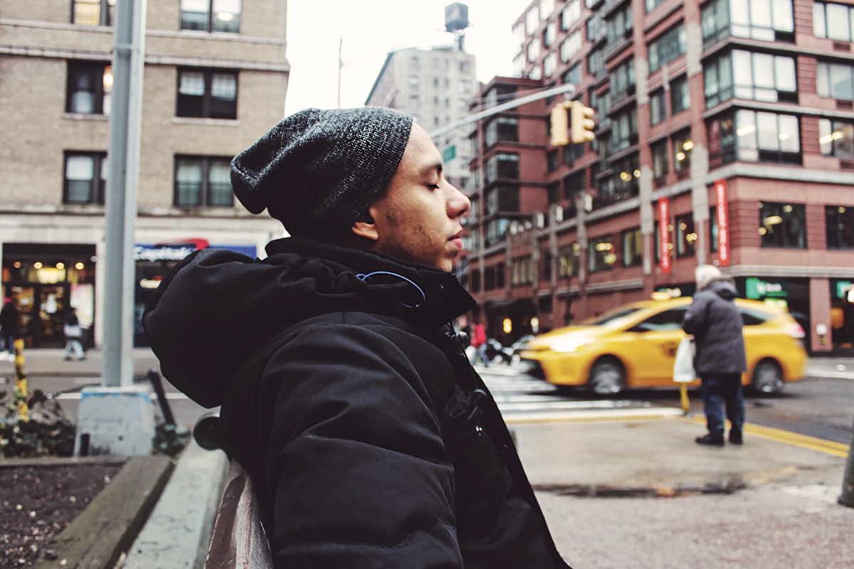 A photo of a young person wearing a black coat and black hat closing their eyes on a busy city street.