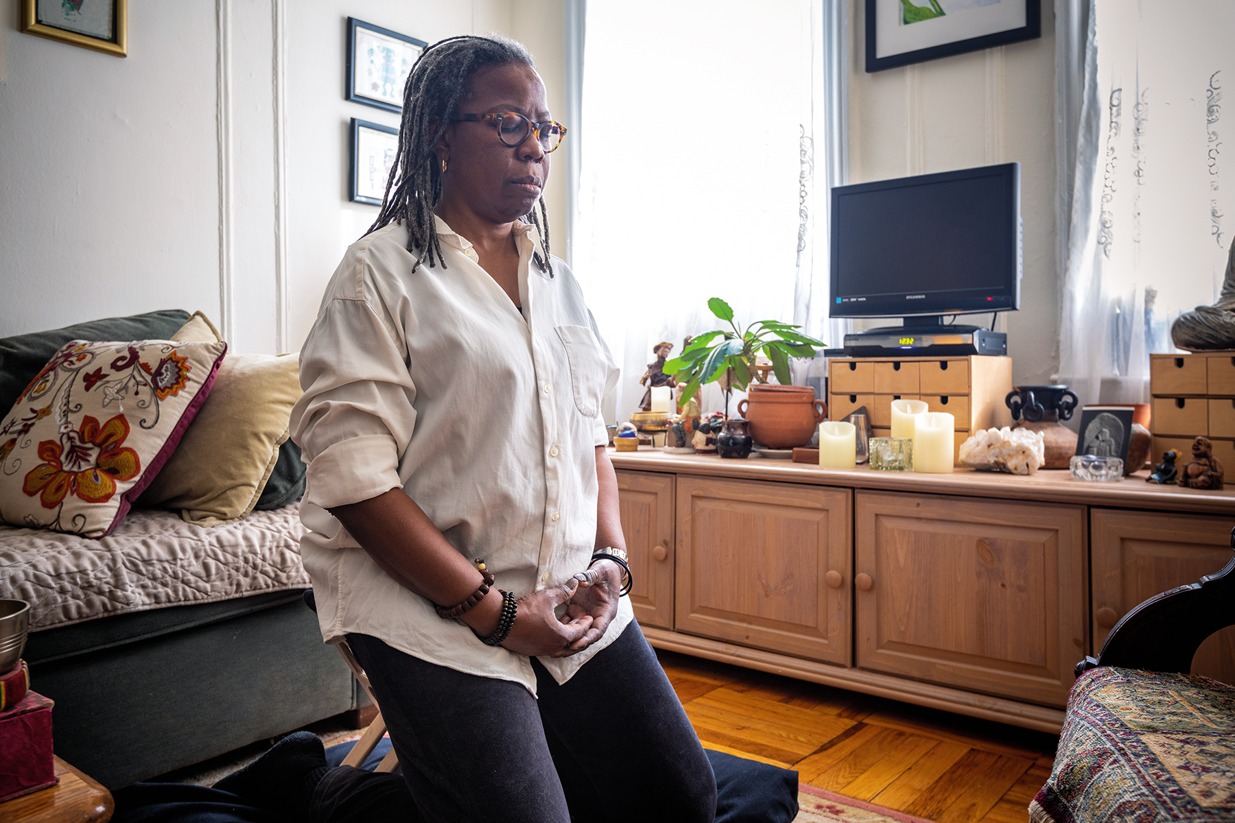 A woman meditating in her house. She is wearing a white shirt and black pants. Her eyes are closed.