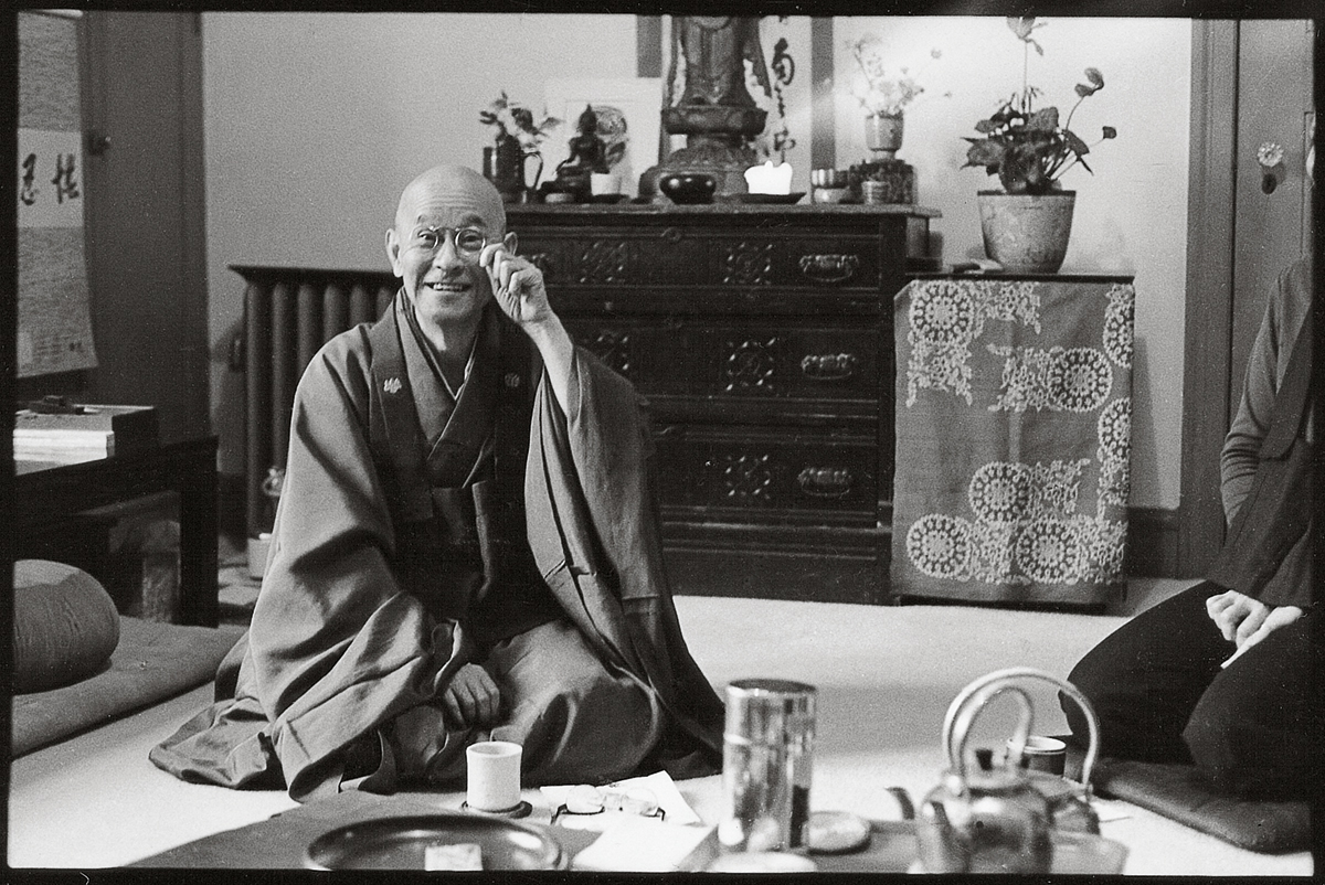 A person sitting on the ground in robes. They are fixing their circle shaped glasses on their face.