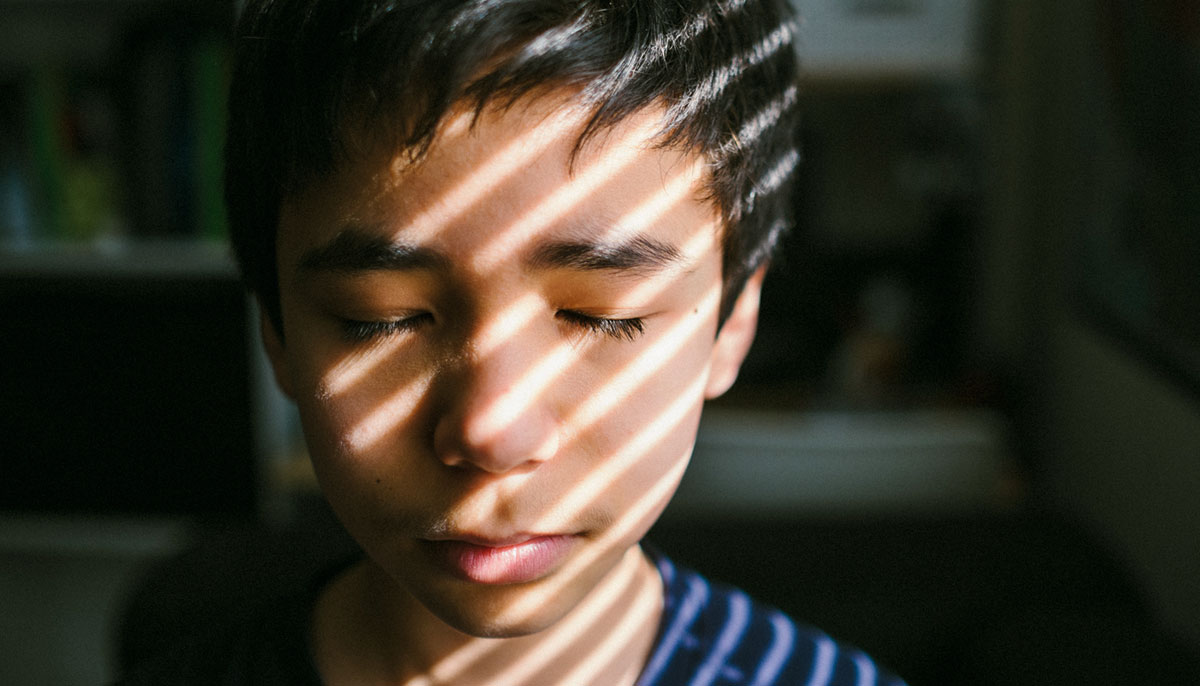 A young boy stands with his eyes closed, the lights of a blind peeking through the window.