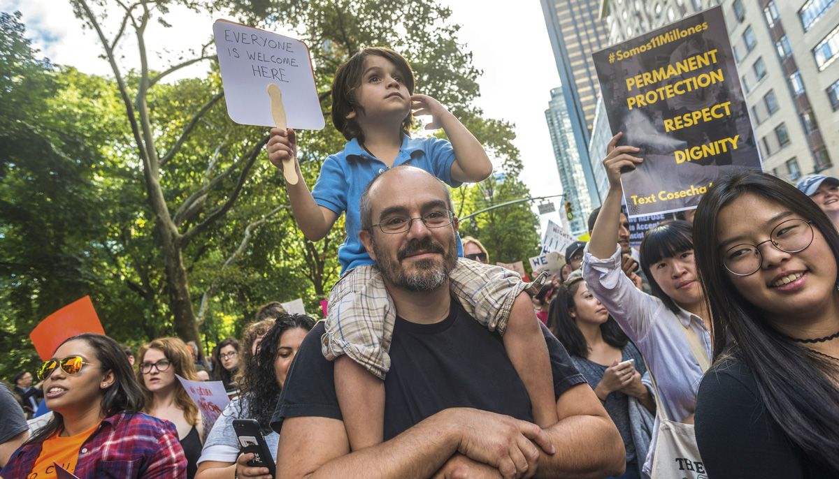 A man with a young child sitting on his shoulders with a sign that reads "everyone is welcome here." They are in a crowded protest.