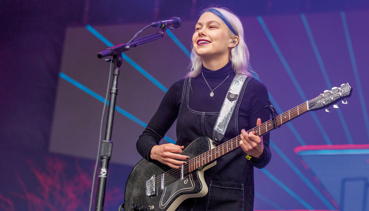 A picture of a woman performing with an electric guitar in front of a microphone. She has a blue streak in her hair and is wearing overalls.